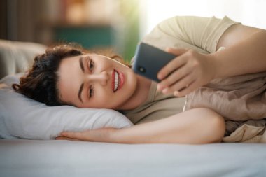 Mutlu, sıradan, güzel bir kadın evdeki yatakta telefon kullanıyor..