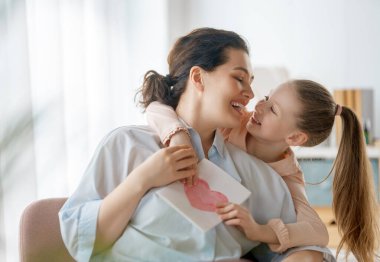 Anneler Günün kutlu olsun. Çocuk kızı annemi tebrik ediyor ve ona kartpostal veriyor. Anne ve kız gülümsüyor ve sarılıyorlar. Aile tatili ve birliktelik.