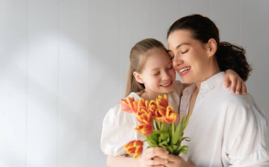Anneler Günün kutlu olsun! Çocuk kızı annemi tebrik ediyor ve ona çiçek laleleri veriyor. Anne ve kız beyaz arka planda gülümsüyor. Aile tatili ve birliktelik