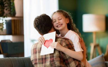 Çocuk kızı babasını tebrik ediyor ve ona kartpostal veriyor. Baba ve kız gülümsüyor ve sarılıyorlar. Aile tatili ve birliktelik.