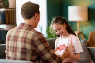 Çocuk kızı babasını tebrik ediyor ve ona kartpostal veriyor. Baba ve kız gülümsüyor. Aile tatili ve birliktelik.
