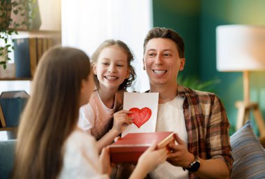 Çocuklar babalarını tebrik ediyor ve ona kartpostal ve hediye veriyorlar. Baba ve kızlar gülümsüyor ve sarılıyorlar. Aile tatili ve birliktelik.