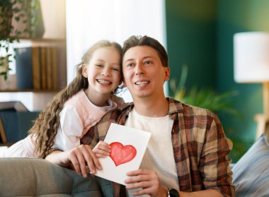 Çocuk kızı babasını tebrik ediyor ve ona kartpostal veriyor. Baba ve kız gülümsüyor ve sarılıyorlar. Aile tatili ve birliktelik.