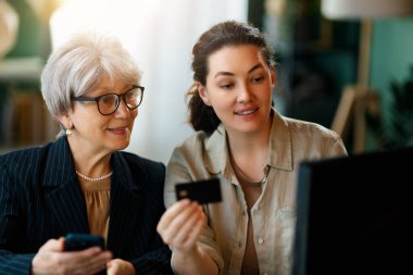 Genç ve yaşlı kadınlar online alışveriş için bilgisayar kullanıyorlar. Kızı evde annesine yardım ediyor..