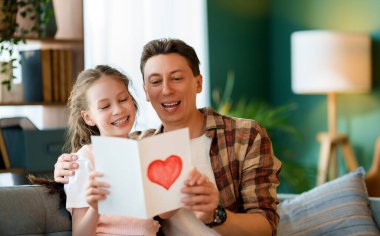 Çocuk kızı babasını tebrik ediyor ve ona kartpostal veriyor. Baba ve kız gülümsüyor ve sarılıyorlar. Aile tatili ve birliktelik.