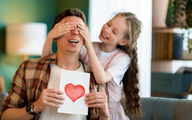 Çocuk kızı babasını tebrik ediyor ve ona kartpostal veriyor. Baba ve kız gülümsüyor ve sarılıyorlar. Aile tatili ve birliktelik.