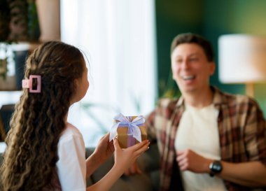 Çocuk kızı babasını tebrik ediyor ve ona bir hediye veriyor. Baba ve kız gülümsüyor. Aile tatili ve birliktelik.