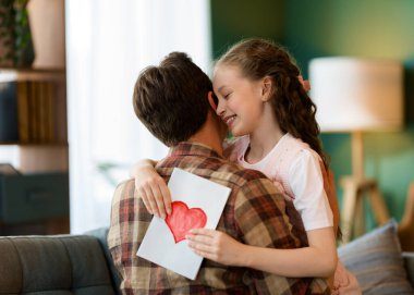 Çocuk kızı babasını tebrik ediyor ve ona kartpostal veriyor. Baba ve kız gülümsüyor ve sarılıyorlar. Aile tatili ve birliktelik.