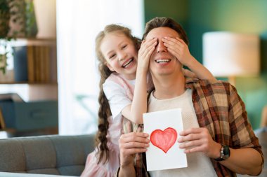 Çocuk kızı babasını tebrik ediyor ve ona kartpostal veriyor. Baba ve kız gülümsüyor ve sarılıyorlar. Aile tatili ve birliktelik.