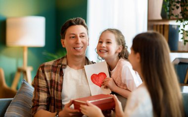 Çocuklar babalarını tebrik ediyor ve ona kartpostal ve hediye veriyorlar. Baba ve kızlar gülümsüyor ve sarılıyorlar. Aile tatili ve birliktelik.