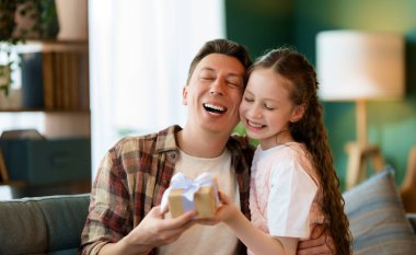 Çocuk kızı babasını tebrik ediyor ve ona bir hediye veriyor. Baba ve kız gülümsüyor. Aile tatili ve birliktelik.