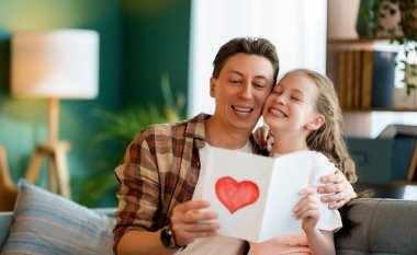 Çocuk kızı babasını tebrik ediyor ve ona kartpostal veriyor. Baba ve kız gülümsüyor ve sarılıyorlar. Aile tatili ve birliktelik.