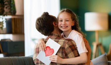 Çocuk kızı babasını tebrik ediyor ve ona kartpostal veriyor. Baba ve kız gülümsüyor ve sarılıyorlar. Aile tatili ve birliktelik.