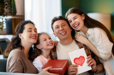 Çocuklar babalarını kutluyor ve ona hediye veriyorlar. Baba, annem ve kızlar gülümsüyor ve sarılıyorlar. Aile tatili ve birliktelik.