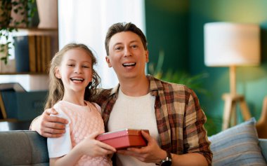 Çocuk kızı babasını tebrik ediyor ve ona bir hediye veriyor. Baba ve kız gülümsüyor. Aile tatili ve birliktelik.