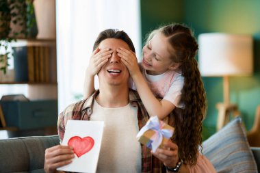 Çocuk kızı babasını tebrik ediyor ve ona kartpostal veriyor. Baba ve kız gülümsüyor ve sarılıyorlar. Aile tatili ve birliktelik.