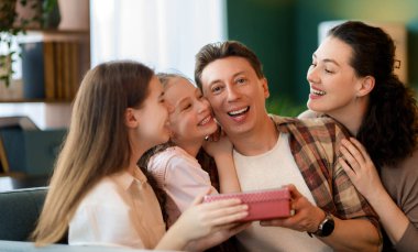 Çocuklar babalarını kutluyor ve ona hediye veriyorlar. Baba, annem ve kızlar gülümsüyor ve sarılıyorlar. Aile tatili ve birliktelik.