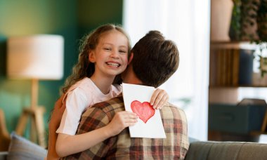 Çocuk kızı babasını tebrik ediyor ve ona kartpostal veriyor. Baba ve kız gülümsüyor ve sarılıyorlar. Aile tatili ve birliktelik.