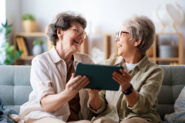 Mutlu yaşlı kadınlar evdeki rahat koltukta otururken dijital tablet kullanıyorlar..