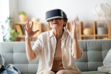 Evde VR gözlük takan yaşlı bir kadın konsol oyunu oynuyor..