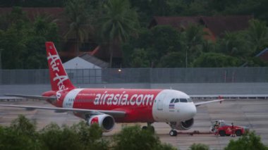 PHUKET, THAILAND - NOVEMBER 28, 2019: Tractor towing Airbus A320 of AirAsia at Phuket airport. Airport on tropical island, airfield and palm trees. Tourism and travel concept