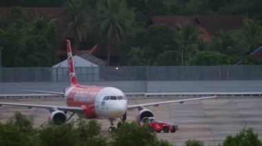 PHUKET, THAILAND - NOVEMBER 28, 2019: Tractor towing plane of AirAsia at Phuket airport. Airport on tropical island, airfield and palm trees. Tourism and travel concept