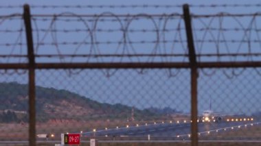 Uçak hızlanıyor ve kalkıyor, havaalanı çitlerinden bakıyor. İniş ışıklarıyla podyum ışıkları