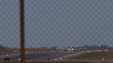 Tanımlanamayan bir üniforması olan özel bir jet alacakaranlıkta havalanmak için hızlanır. Havaalanı çitlerinden bak. İniş ışıkları, ışıklandırma, pist