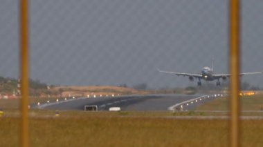 Tanımlanamayan bir üniforması olan, iniş, dokunma ve frenleme yapan bir uçağın yavaş çekim görüntüleri. Havaalanı çitlerinden geçen pistin görüntüsü. İniş ışıklarıyla aydınlatılmış iniş pisti