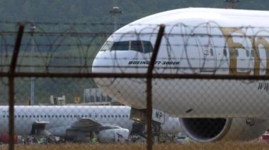 PHUKET, THAILAND - 25 HAZİRAN 2023: Emirates 'ten Boeing 777 tipi geniş gövdeli uçak Phuket Havaalanı, yan görüş pistinde taksicilik yapıyor. Havaalanı çitlerinin dışındaki taksideki uçak. Seyahat kavramı