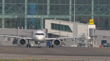 Önlükteki jet uçağının ön görüntüsü. Uçak taksicilik yapıyor. Arkaplan terminali, hava köprüsü, kol