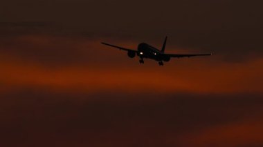 Jet uçağı gün batımında. Uçak uçuyor, alacakaranlıkta inişe geçiyor. Koyu kırmızı gökyüzü ve uçağın silüeti.