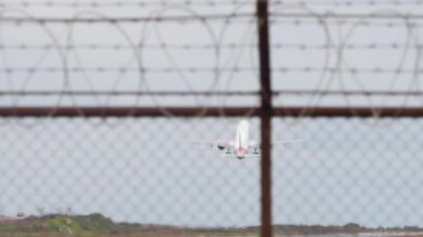 Ticari bir jet gökyüzüne yükseliyor, tel örgülü bir havaalanı çitinden bakıyor. Karamsar ve dinamik sahne seyahat, kalkış ve modern hava taşımacılığı kavramları için mükemmeldir..