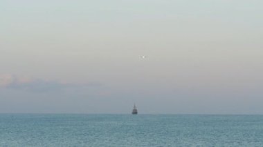 Ufukta görünen büyük bir gemiyle denizin üzerine inen bir jet uçağının uzaktan görüntüsü..
