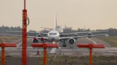 İki ticari uçak pistte sırayla hareket ediyor, ön taraftan görülüyor. Uçaklar yavaş hareket ediyor, kalkışa hazırlanıyorlar. Hava yolculuğu temaları için uygun