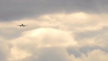 Bulutlu gökyüzüne iniş için yaklaşan ticari bir uçağın uzaktan görüntüsü. Yolcu jeti piste doğru iniyor, son yaklaşımı yakalıyor. Havacılık, seyahat ve hava taşımacılığı.