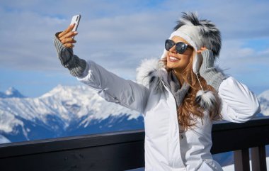 Teknoloji ve eğlence kavramı. Kış kürkü şapkalı mutlu kadın dışarıda akıllı telefonuyla selfie çekiyor.                      
