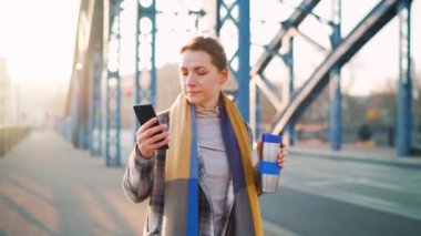 Beyaz ceketli bir iş kadını, soğuk güneşli bir sabahta şehirde dolanıyor, kahve içiyor ve akıllı telefon kullanıyor. İletişim, iş günü, yoğun yaşam konsepti.