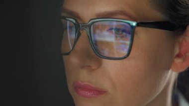 Woman in glasses looking on the monitor and surfing Internet at night. The monitor screen is reflected in the glasses. Work at night. Remote work. Extreme close-up