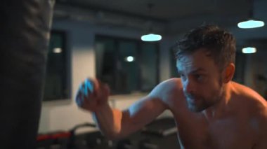 Alone male boxer is working out blows on a punching bag in a gym in evening. Slow motion