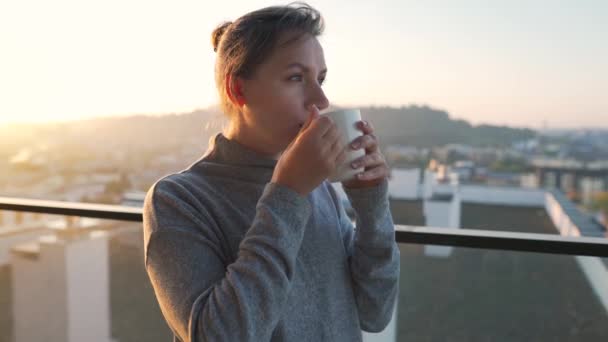 女人在黎明时分在阳台上喝杯茶或咖啡开始她的一天 动作缓慢 以城市景观为背景 现代城市生活方式 — 图库视频影像
