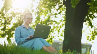 Günbatımında şehir parkında çimlerde oturan laptopta çalışan çekici bir kadın. Dışarıda çalışan serbest çalışan biri.