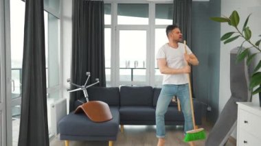 Adam evi temizliyor, dans ediyor ve süpürgeyle şarkı söylüyor. Yavaş çekim. 