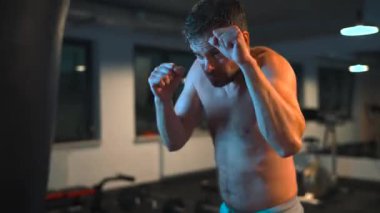 Alone male boxer is working out blows on a punching bag in a gym in evening.