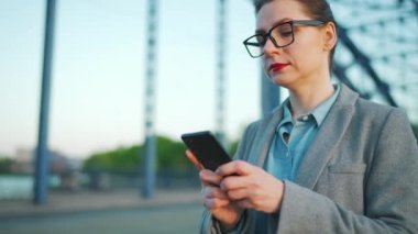 Sabahın erken saatlerinde şehirde dolaşan ve akıllı telefon kullanan beyaz bir iş kadını. İletişim, iş günü, yoğun yaşam konsepti. Yavaş çekim