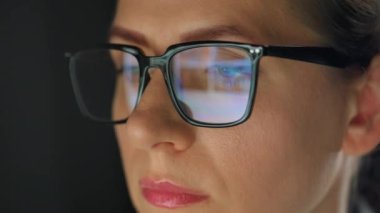 Woman in glasses looking on the monitor and surfing Internet at night. The monitor screen is reflected in the glasses. Work at night. Remote work. Extreme close-up