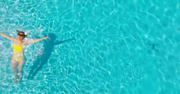 一个穿着黄色泳衣的女人躺在游泳池里的自上而下的照片 照相机上升 顺时针旋转 放松概念 夏季生活方式 — 图库视频影像