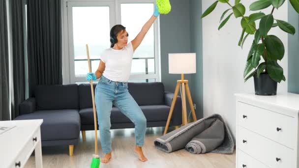 ヘッドフォンで白人女性は家を掃除し ほうきや洗濯で楽しいダンスをしています スローモーション — ストック動画