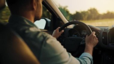Adam yol boyunca araba sürüyor, batan güneş ön camda parlıyor. Sürücülerin omzunun arkasından bak.
