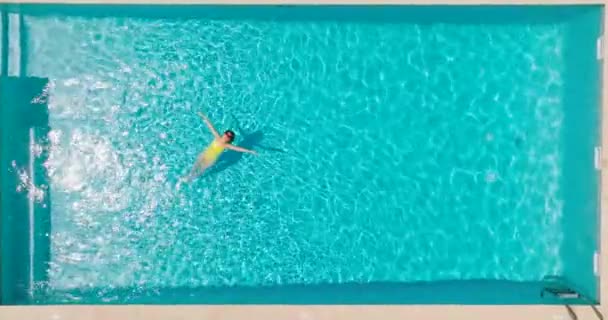 黄色の水着を着た女性のトップダウンビューは プールに彼女の背中に横たわっている リラックスしたコンセプト 夏のライフスタイル カメラが水面に落ちている — ストック動画